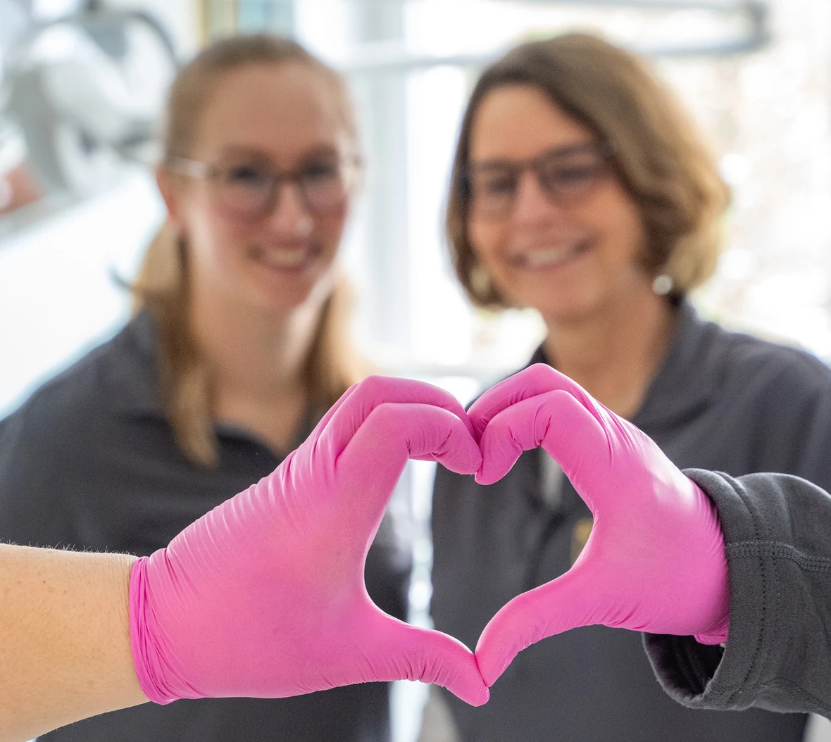 Zwei Kolleginnen der Kieferorthopädie Am Silberkamp formen mit ihren Handschuhen ein Herz